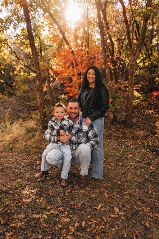 A man crouching on leaf-covered ground with a young boy sitting on his knee, and a woman standing behind them with her hand on his shoulder, in autumn woodland with orange leaves and low sun through the trees